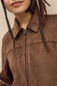Tuvumā redzams cilvēks ar brūnu zamša jaku ar rāvējslēdzēju, redzama daļa no viņa mutes un daži dreadlocki.