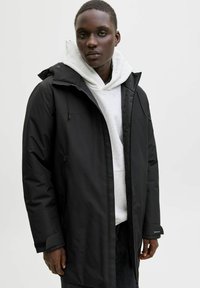 Črni vodoodporni parka s prilagodljivo kapuco, zadrgo in stranskimi žepi. Oblečen čez sivo kapuco, strukturirane tkanine in dolge rokave.