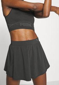 Musta urheilullinen lyhyt toppi, jossa on Puma-logo, yhdistetty mustaan liehukehameeseen. Kangas on sileä ja siinä on hieman kiiltoa sekä joustava vyötärö.