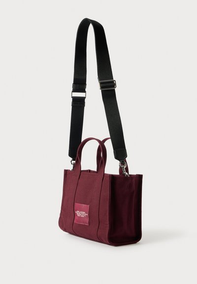 Sac fourre-tout en toile bordeaux avec deux poignées supérieures, bandoulière noire réglable et une petite étiquette rose avec du texte blanc, design minimal.