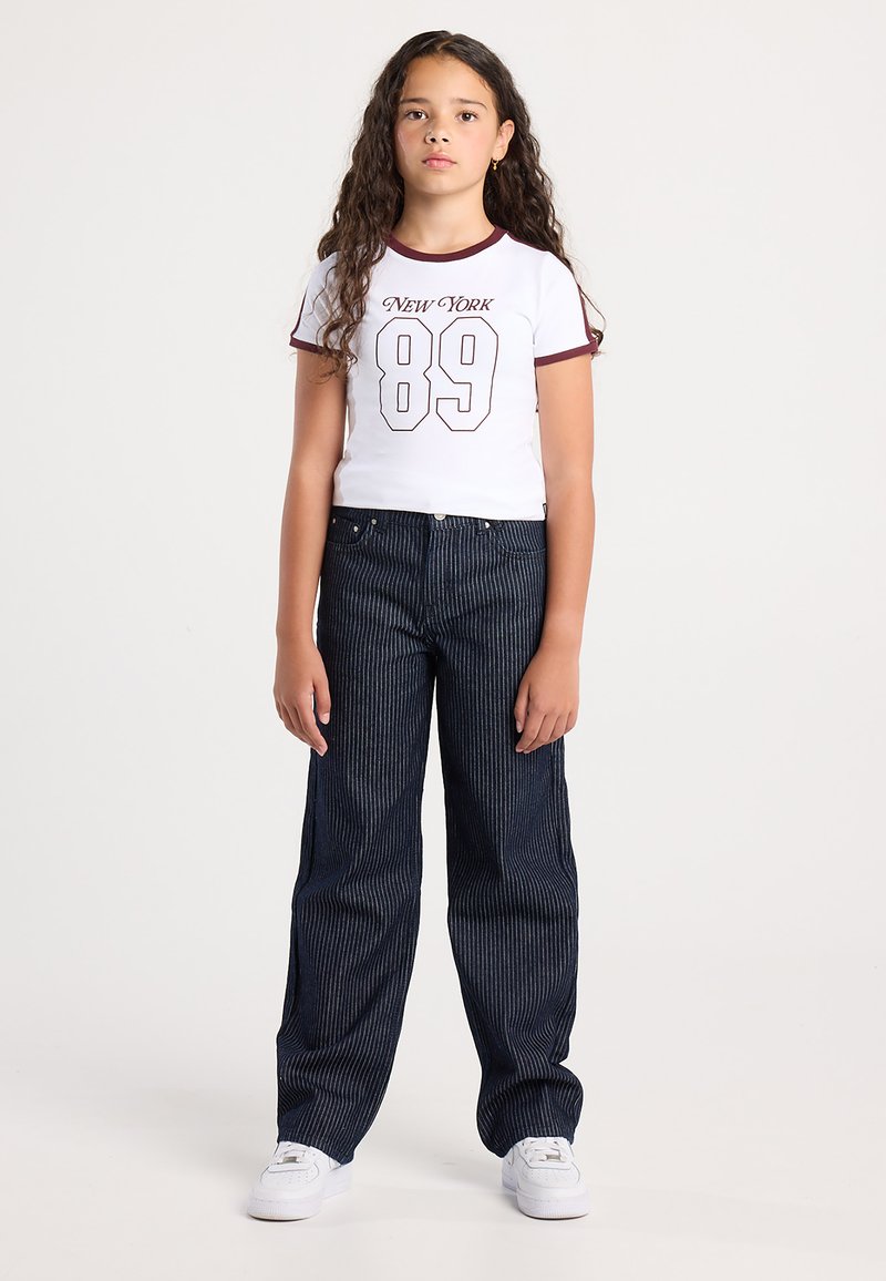 Ragazza con lunghi capelli ricci che indossa una maglietta bianca "New York 89", pantaloni larghi a righe scure e sneakers bianche, in piedi davanti a uno sfondo semplice.