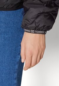 Black puffer jacket sleeve with quilted texture, featuring a white logo band with "TOMMY JEANS." Worn over blue denim with a close-up of the hand.