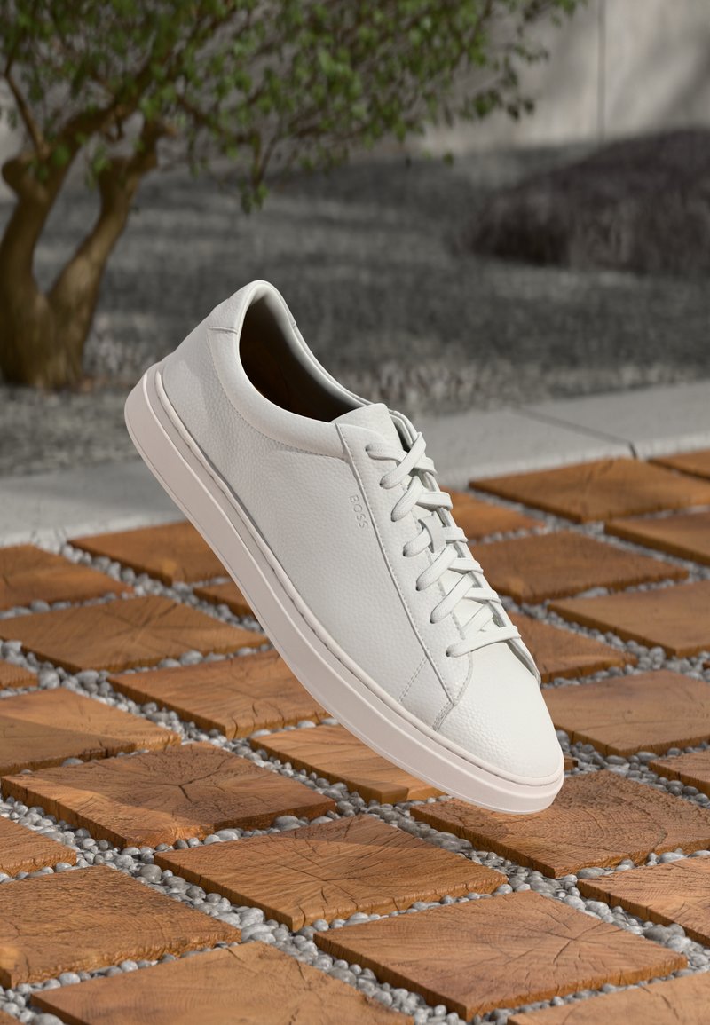 Weiße Leder-Sneaker mit strukturierter Oberfläche, runder Zehenpartie und flachen Schnürsenkeln, vor einem Holztile-Hintergrund mit Kieselsteinen.