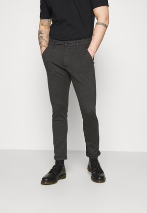 Mann trägt dunkelgraue Slim-Fit-Hose, schwarze Socken, schwarze Leder-Schnürschuhe und ein schwarzes T-Shirt, wobei tätowierte Arme sichtbar sind.