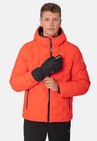 Leuchtend orangefarbene Daunenjacke mit Reißverschluss und Kapuze. Mit schwarzem Reißverschluss und Akzenten. Das Modell trägt schwarze Handschuhe. Glatte Textur.