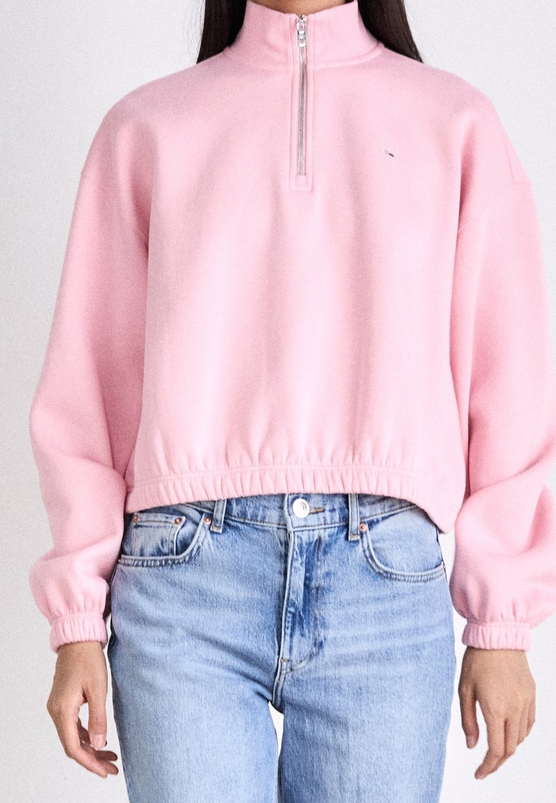 Felpa rosa con collo a zip, vestibilità rilassata, polsini e orlo a coste e texture liscia, abbinata a jeans in denim azzurro.