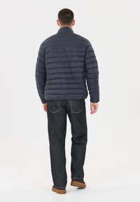 Mörkblå pufferjacka med horisontell sömnad, ståkrage och långa ärmar. Bärs med mörka jeans och bruna skor.