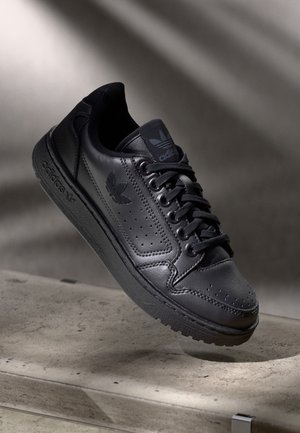 Sort læder Adidas sneaker med snørebånd, perforerede sider og mærket sål, vist mod en neutral baggrund på en betongulv.