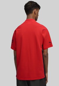 Polo de manga corta rojo con tejido texturizado, con un acento en navy en el costado. Logotipo en la manga izquierda. Color estándar.
