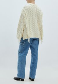 Cremfarbener Kabelstrickpullover mit Fransen, kombiniert mit hellblauen weiten Jeans und schwarzen Stiefeletten mit Absatz.