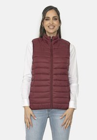 Chaleco acolchado burdeos con cuello alto, cremallera frontal y patrón de acolchado vertical, llevado sobre una camisa blanca de manga larga.