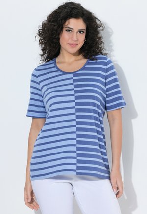 Kvinde med krøllet mørkt hår iført en blå kortærmet skjorte med vandrette striber i halvlyse og halvmørke nuancer og hvide bukser.