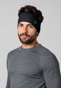 Bandeau noir avec des détails de couture et un logo sur le côté, porté par un homme en chemise grise à manches longues. Texture du tissu lisse.