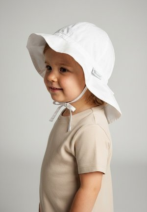 Kleines Kind in beigem Hemd mit weißem Sonnenhut mit Kinnriemen, lächelt und schaut nach links vor einfachem hellem Hintergrund.