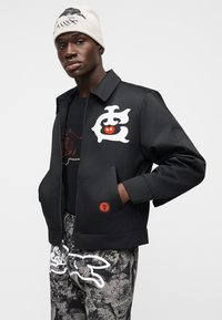 Μαύρο μπουφάν με ψηλό γιακά, μεγάλο λευκό γραφικό, πλαϊνές τσέπες και κόκκινη λεπτομέρεια. Φορεμένο πάνω από μαύρο T-shirt και παντελόνι με σχέδια.