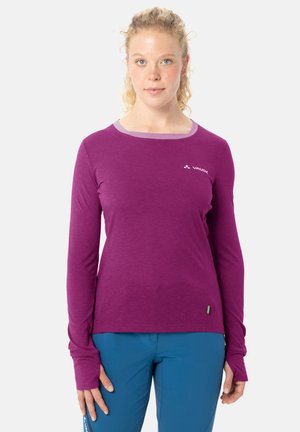 Vrouw met krullend blond haar, gekleed in een paarse lange-mouwen Vaude-trui en blauwe broek, staand tegen een witte achtergrond.