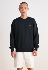 Schwarzes Sweatshirt mit Rundhalsausschnitt und langen Ärmeln, das ein weißes Jumpman-Logo auf der Brust trägt. Kombiniert mit weißen Shorts.