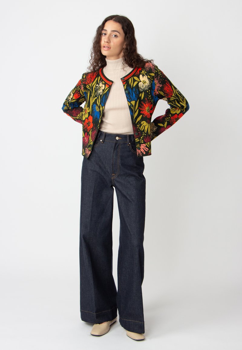 Veste brodée florale colorée sur un fond noir, assortie à un pantalon en denim sombre taille haute et un haut en col roulé clair.