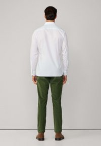 Man staat met zijn rug naar de camera in een witte lange mouw shirt, groene corduroy broek en bruine schoenen tegen een effen achtergrond.