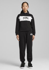 Sudadera negra con una franja horizontal blanca que presenta el logo de "PUMA", combinada con pantalones jogger negros, ambos hechos de tejido suave. Zapatillas negras.