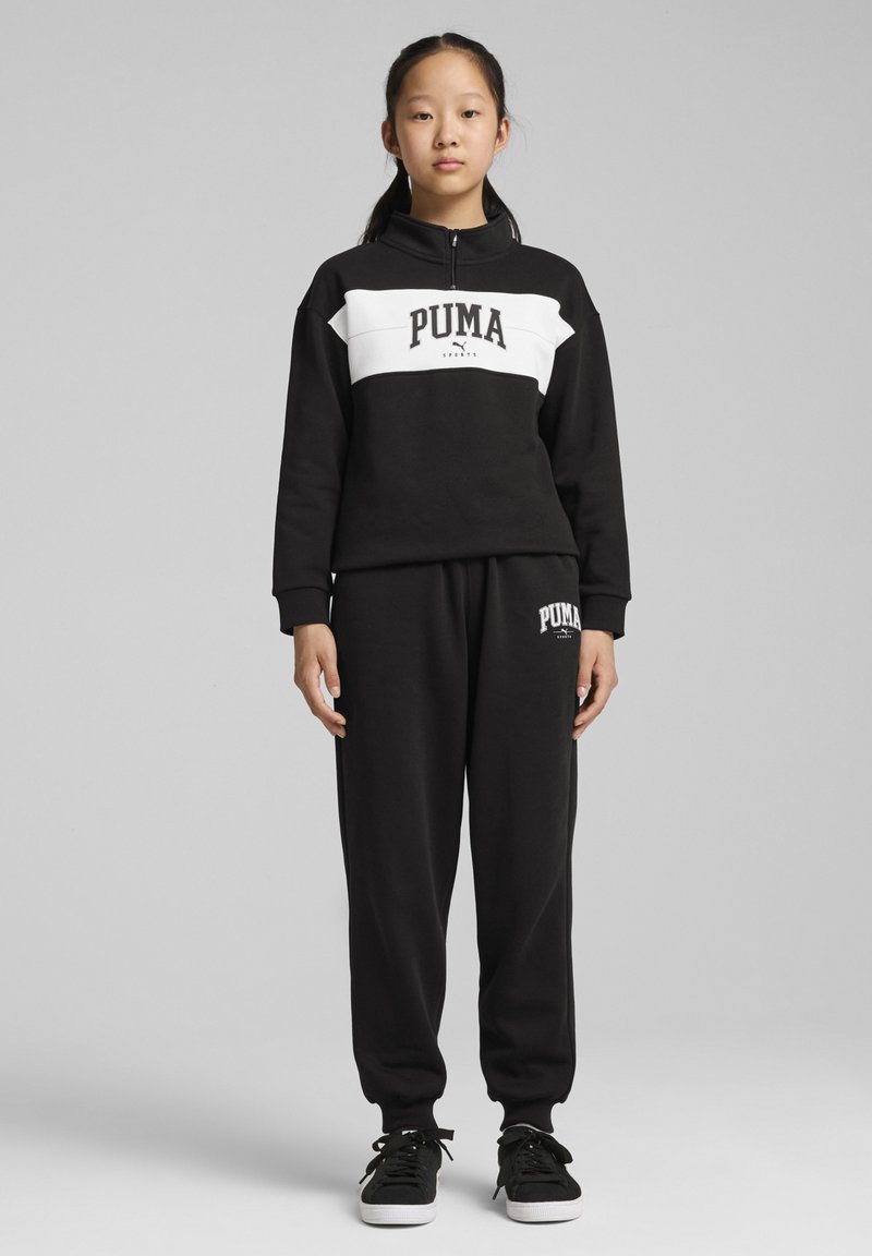 Sudadera negra con una franja horizontal blanca que presenta el logo de "PUMA", combinada con pantalones jogger negros, ambos hechos de tejido suave. Zapatillas negras.