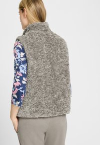 Grijze, fluffy vest met een hoge kraag; heeft een zachte textuur. Draagt over een gemusterd, langemouwen shirt, gecombineerd met grijze broek.