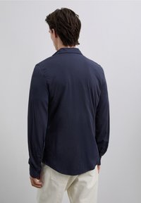 Navy blue long-sleeve shirt, smooth texture, buttoned cuffs, notch collar, curved hem, paired with light-colored pants.