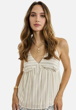 Mujer con cabello largo y ondulado de color castaño, que lleva una blusa sin mangas a rayas beige y blancas, un collar de cadena dorada con un colgante y una pulsera dorada.