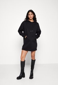 Schwarzes Kapuzen-Sweatshirtkleid mit Kängurutasche, strukturiertem Stoff und gerippten Bündchen. Kombiniert mit kniehohen schwarzen Stiefeln.