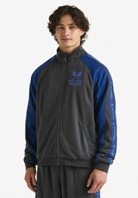 Giacca zip-up grigia con accenti blu. Presenta ricami a tema tennis e dettagli a righe sulle maniche. Realizzata in materiale morbido e liscio.