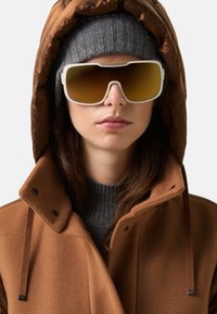 Sluneční brýle s bílým rámem a hnědými skleněnými čočkami, nošené s šedou pletenou čepicí a hnědým péřovým kabátem s vysokým límcem.