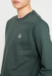 Ciemnozielona bluza z materiału o gładkiej strukturze. Posiada małe białe logo pingwina na piersi oraz ściągacze przy kołnierzu i mankietach.