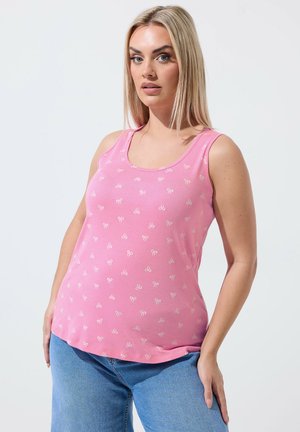 Vrouw met lang blond haar, gekleed in een roze mouwloos topje met kleine witte strikjes en een blauwe spijkerbroek, staand tegen een effen achtergrond.