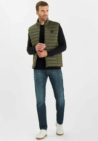 Olivengrön vadderad väst med hög krage, i kombination med en svart långärmad skjorta och blå jeans, kompletterat med vita sneakers.