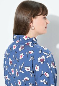 Blaues Blusenshirt mit hohem Kragen, das weiße Blumen und rote Akzente aufweist. Glatter Stoff mit tailliertem Schnitt und langen Ärmeln.