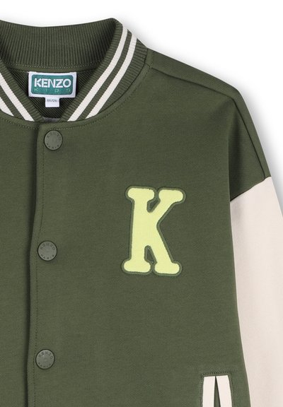 Veste en jersey vert avec col rayé, présentant une grande lettre "K" jaune appliquée, des boutons noirs et des manches de couleur crème.