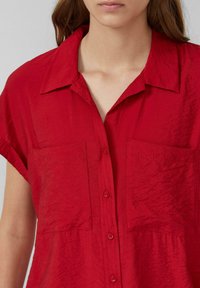 Rotes Kurzarmhemd aus strukturiertem Stoff. Mit Knopfverschluss, Kragen und zwei Brusttaschen. Glatte Oberfläche mit zerknittertem Aussehen.