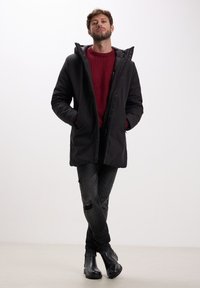 Parka nera isolata con cappuccio, caratterizzata da una chiusura con zip, tasche laterali e un maglione rosso a trama sotto. Indossata con jeans neri strappati e stivali neri.