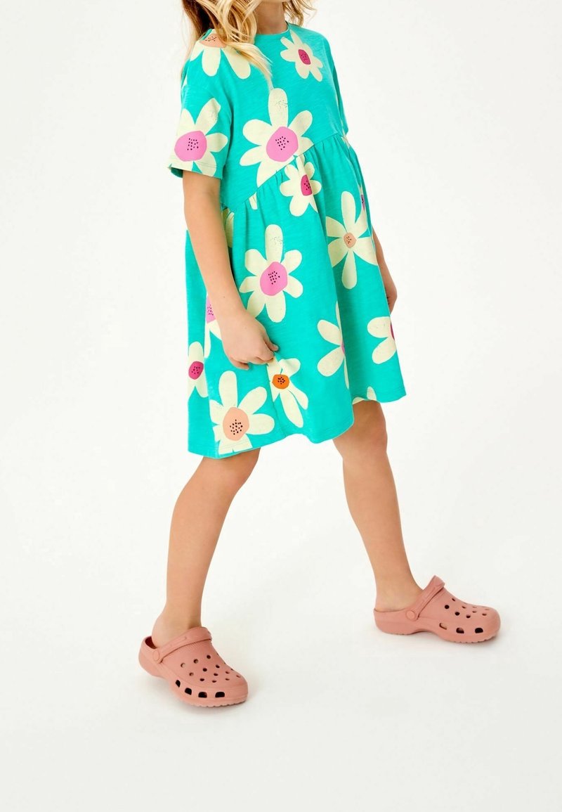 Enfant portant une robe turquoise à grandes fleurs crème et rose, avec des chaussures légères roses, debout devant un fond blanc uni.