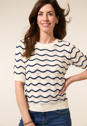 Lachende vrouw die een wit kort mouwen trui met marineblauwe golvende strepen draagt, blauwe jeans, gouden oorbellen, armband, en met haar hand op haar heup tegen een lichte achtergrond.