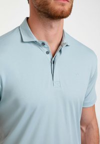 Šviesiai mėlynas polo marškinėlis, pagamintas iš minkštos medžiagos, su apykakle, dviem mygtukais ir mažu logotipu ant krūtinės. Trumpomis rankovėmis ir lygiu paviršiumi.