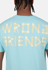 Camiseta de algodón azul claro con la frase "WRONG FRIENDS" en letras de cinta beige en la parte posterior. Diseño de manga corta y cuello redondo.