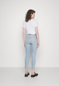 Jeans de cintura alta azul claro com corte slim, bainhas desfiadas e bolsos traseiros clássicos, combinados com uma camisa branca de mangas curtas e sapatos pretos.