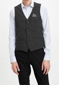 Uomo che indossa un gilet grigio antracite con una pochette a fantasia sopra una camicia da dress blu chiaro e pantaloni neri.