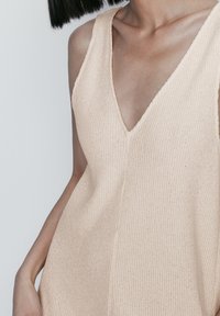 Beige gestricktes ärmelloses Oberteil mit V-Ausschnitt, das eine gerippte Textur und einen lockeren Schnitt aufweist. Dezente Nähte entlang der Mittelnaht.