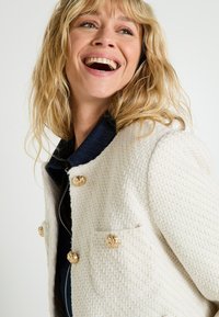 Chaqueta blanca texturizada con acentos de botones dorados, que presenta un cuello de mezclilla azul y un cierre de cremallera. La tela tiene un patrón tejido.