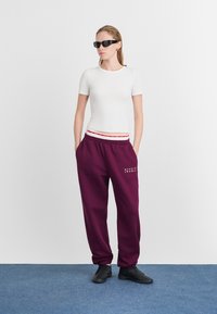 Bijela prilagođena majica, bordo trenirke s crvenim i bijelim pojasom, detalj s logom i crne atletske tenisice. Model nosi crne sunčane naočale.