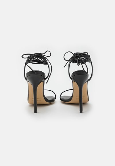 Sandalias negras de tacón alto con tiras delgadas y lazos en la parte posterior. Presentan un elegante tacón de aguja y un acabado suave y brillante.