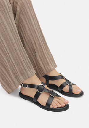 Nohy v černých kožených sandálech se zlatými kroužky a béžové široké kalhoty s žebrovaným vzorem na bílém pozadí.