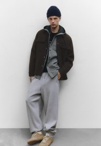 Outfit stratificato con giacca marrone scuro, pantaloni della tuta grigi oversize e camicia a quadri. Il look è completato da un berretto scuro.